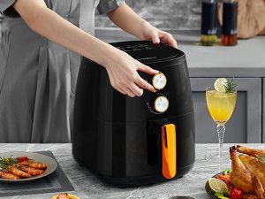 Air Fryer: A person adjusting the dials on a black air fryer in a modern kitchen, with an air fryer-prepared plated shrimp dish and air fryer-roasted chicken on the countertop, alongside a glass of lemonade.