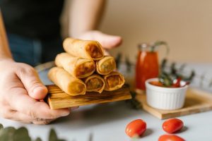 Air Fryer: A person's hands presenting a stack of golden-brown spring rolls made in an air fryer, displayed on a wooden board with fresh tomatoes and a small bowl of dipping sauce in the background.