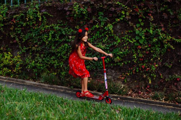A pretty little girl in a flowery dress and with a cute red headband is riding her scooter without her parents supervision. 