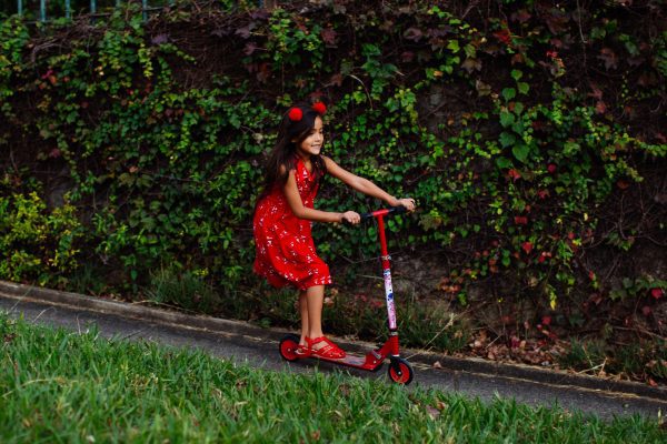 A pretty little girl in a flowery dress and with a cute red headband is riding her scooter without her parents supervision.