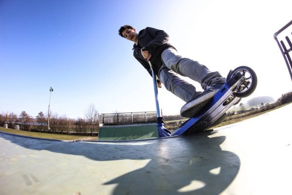 A man wearing black long sleeve is riding his scooter with a new applied deck tape at the scooter park on a bright and sunny morning.