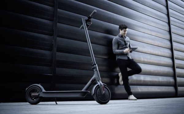 A man is leaning on the wall with its black scooter with new applied deck tape. Its scooter is elegant due to its deck tape.