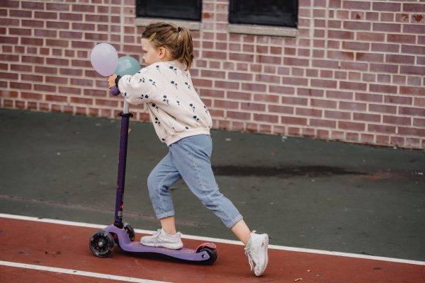 A little girl riding her purple scooter on a red track near a building with a wall that is made out of red bricks.