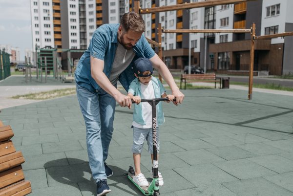 A father in blue polo teaches his child to scoot in an open space away from vehicles and accidents. They are practicing outside in spacious area to scoot freely and safely.