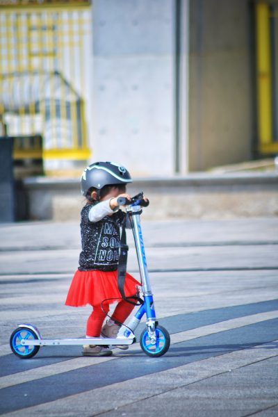 A cute little girl is riding a blue scooter while wearing her helmet, jacket, and red skirt. She is practicing alone without her parents supervising.