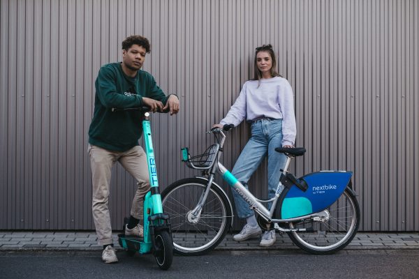 A man is safely posing with his bright colored scooter and a beautiful girl with her bike.