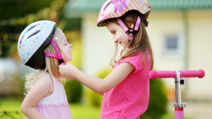 Two kids wearing helping each other wearing a helmet before riding a scooter.