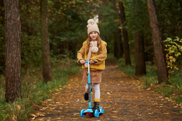 A pretty girl with a yellow jacket is riding her blue scooter in the woods.