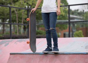 A half body of an adult holding a skateboard. The person is wearing white shirt and denim pants. This picture was taken in skateboard park. 