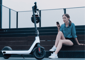 A woman wearing a blue shirt and black shorts sits and checks her phone while resting from her electric scooter ride. 