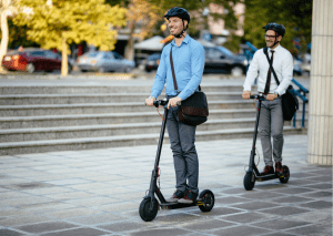 Two helmeted men wearing sling bags smile while each enjoying the best ride on their electric scooter outdoors.