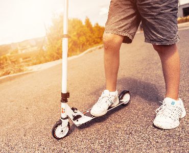 A person wearing white sneakers rides a scooter outdoors on a bright day. 