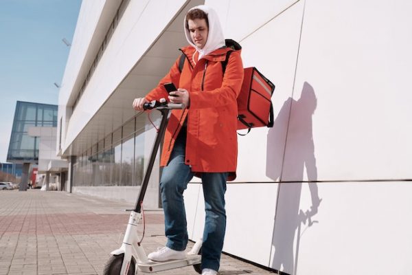 A man in orange checking his phone while standing and holding an electric scooter