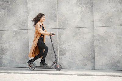 A woman with long curly hair wearing a brown coat strolls using her scooter.