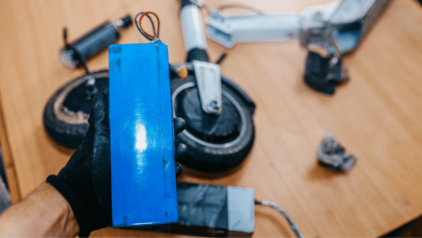 A hand holding a color blue battery. The background picture is a disassembled electric scooter. 