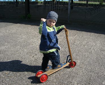 A young kid wearing a bonnet holds the handlebar of his scooter outdoors. 