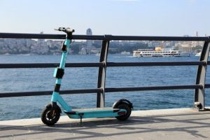 Scooters for kids: a turquoise-framed model stands on a seaside promenade, facing a busy waterway with boats.
