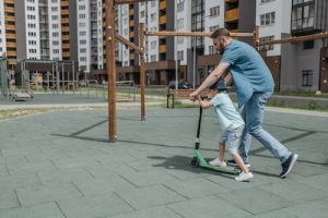 A father teaches his child to ride scooters for kids in a playground setting.