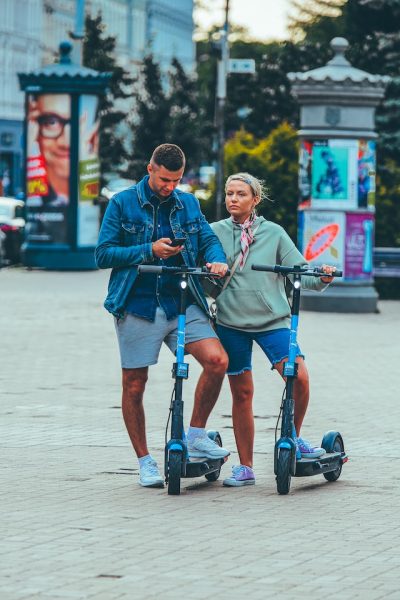 A man and woman of different age standing on their electric scooter as the former checks his mobile phone.