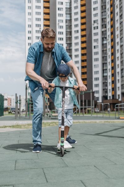 A middle aged dad teaching his son how to ride a scooter. 