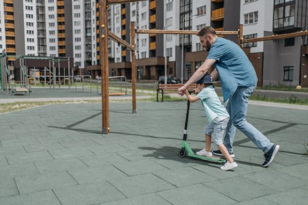 Dad teaching his son to ride a scooter in the park.