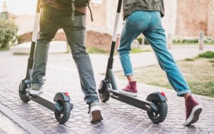 Scooter Deck:The image captures two individuals mid-stride, each with one foot on an electric scooter deck, stepping on the scooter decks, ready to ride along a paved outdoor path. Each scooter deck is black. Each scooter deck is flat.