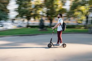 This is a good picture of a man happily strolling with his black electric scooter in the park after his class. This man had an absolutely wonderful day.