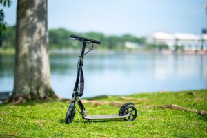 Deck: The image showcases a standalone electric scooter parked on a grassy area beside a tree, with its scooter deck clearly visible. The background features a serene lake, suggesting a peaceful park setting. The scooter deck is sturdy. Each scooter deck is flat.