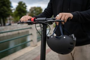 Scooter Deck: Combine fun and safety with your scooter deck. The image displays a close-up of scooter decks with a person holding the handlebars of an electric scooter and a helmet. The scooter deck is black. The scooter deck is flat.