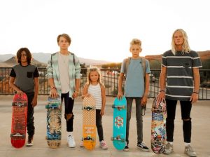 Four aspiring skateboarders of varying ages and heights stand side-by-side against an expansive skatepark backdrop, bathed in the golden glow of the setting sun. Each holds their personal skateboard, displaying a mix of designs and colors, showcasing their individuality. The serene end-of-day light creates long shadows on the ground, highlighting the shared bond and silent anticipation of the young riders before they embark on their escapades. As the day wanes, the calm before the flurry of evening activity is almost tangible, with each mentally preparing for the tricks and rides to come under the fading light.