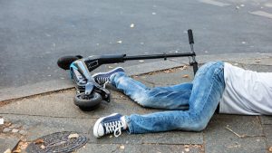 Amidst the urban backdrop, a boy in a white shirt and jeans lies on the street with his scooter, with his half body concealed from view. The scene raises questions about the circumstances that led to his position. 