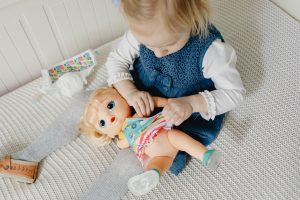 A little girl wearing a blue dress plays with her baby doll inside her baby room. This baby doll can provide many benefits for the little girl. Among the benefits of a baby doll is the development of essential life skills. Engaging in imaginative play with a baby doll also encourages creativity and empathy.