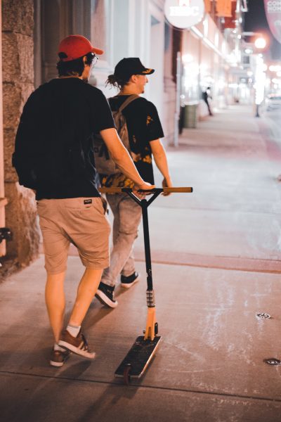 Two men in black shirt with red and black hats and with backpack are walking on the sidewalk with a moped with them 