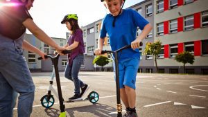 Two kids wearing helmet enjoy their scooters outdoors in front of a big building. 