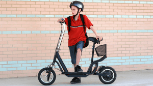 A boy wearing a red shirt and a black helmet rides his scooter with a scooter seat as an added customization. 