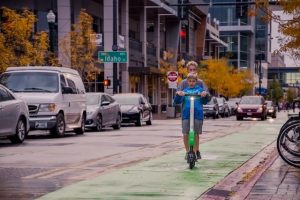 A child rides a scooter with a prominent scooter deck along a green-painted bike lane on a city street. The scooter is white and green.