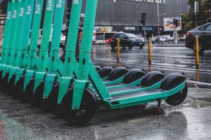 Scooters and Decks: A row of green electric scooters with prominent scooter decks is parked on a wet urban street, glistening with rain. The scooters are arranged neatly, with their handlebars facing upward and wheels touching the ground. Each scooter is the same.