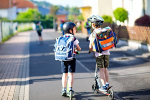 Two school boys are heading home using their kick scooters. They are wearing helmets and talking while riding.