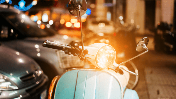 In the picture, there is a scooter with vibrant lights. The scooter's lights are particularly bright, as its headlight is pointing directly at the camera. The photo employs the bokeh effect to create a blurred background, yet it allows us to distinguish other scooters and cars behind the main one. This blue-green scooter stands out, featuring wide handlebars and a high side mirror adorned with additional lights.