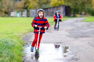 Two little boys riding on kick scooters on the way to or from school. The boys are having fun running their scooters through puddles. 