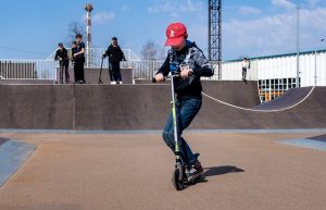 A boy wearing a red cap and long sleeve shirt enjoys practicing tricks in the spacious skate park on a bright day. 