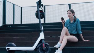 A joyful woman sits comfortably next to her scooter. With her phone in hand, demonstrating the scooter's waterproof capabilities. Waterproof features contribute to an easy maintenance.