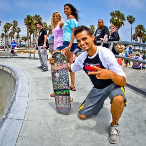 A guy is posing with his board. There are other people in the background who seem looking at the camera.