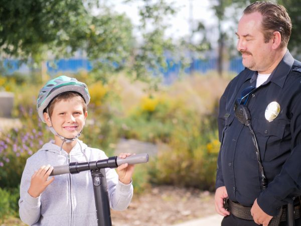 A boy happily using his scooter in the park smiles towards the camera while the guard looks at him gladly. 