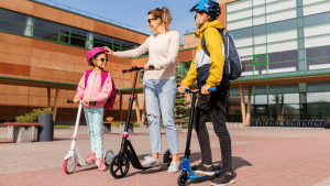 A mom teaching two young kids (around 7-year-old to 10-year-old kids) how to ride a scooter. The kids are wearing blue and pink helmet that matches their scooter. 