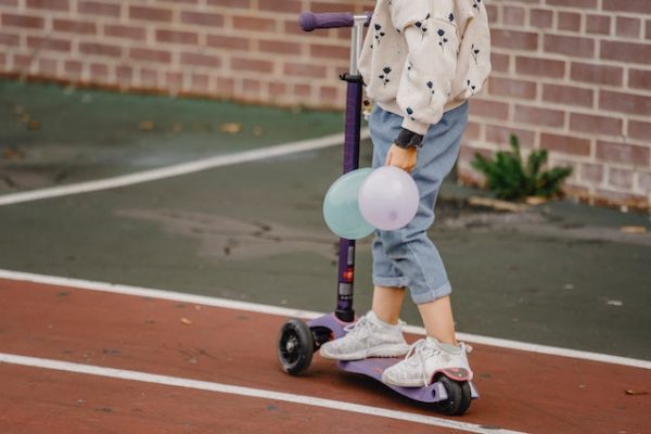 A child riding a scooter while holding balloons