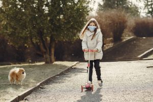 A girl wearing a mask rides her scooter while enjoying the park. 