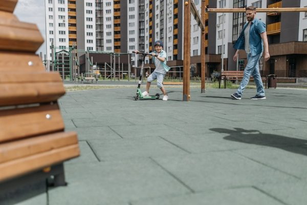 A boy and his dad strolling around the park with the child's scooter. 