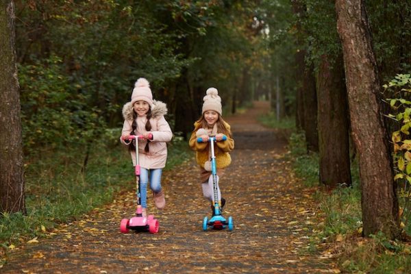 Two girls wearing winter clothes while riding their scooters in a pathway surrounded with trees and plants.