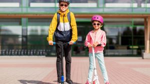 two kids stand side by side, beaming with smiles, as they proudly display their scooters. Both children are responsibly geared up in appropriate safety gear, including helmets, knee pads, and elbow pads. Their beaming faces reflect the joy and excitement of their shared scooter adventure. Amidst the sunlit park, the two kids effortlessly glide on their scooters, their balance and coordination making it seem like second nature